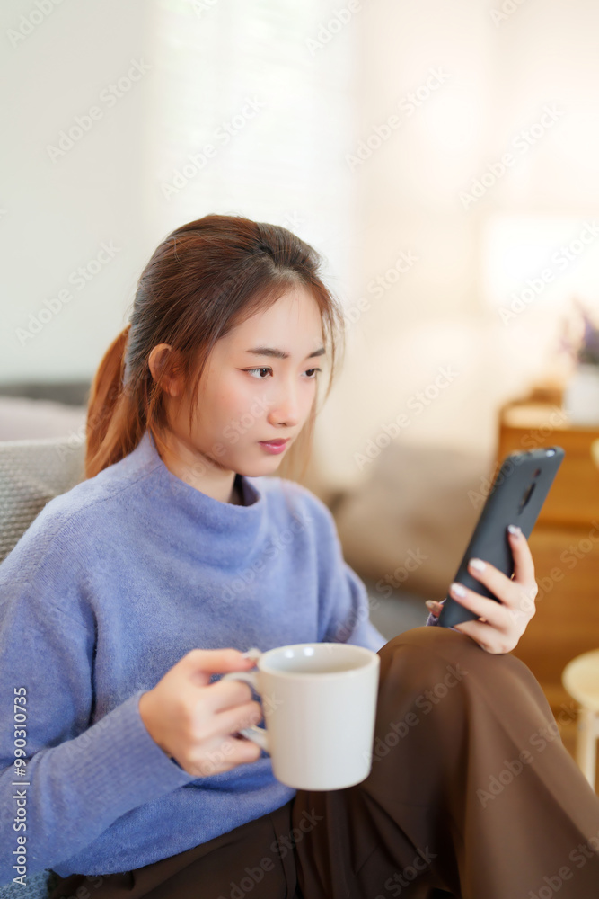 Young asian women in sweater surfing social media on smartphone and drinking coffee while sitting on the couch to relaxation and doing activity for spending time with slow life lifestyle at home Young asian women in sweater surfing social media on smartphone and drinking coffee while sitting on the couch to relaxation and doing activity for spending time with slow life lifestyle at home