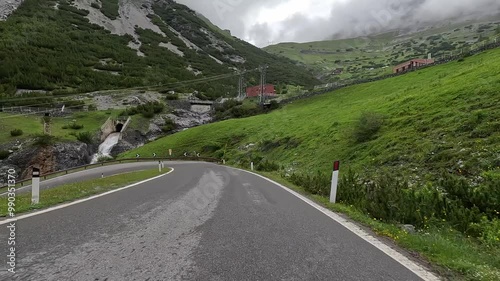 Cyclist descending Stelvio Pass, riding past low trees, green grass, mountain stream, deep valleys with stone cliffs partially shrouded in clouds. Stunning landscape offers challenging and scenic ride