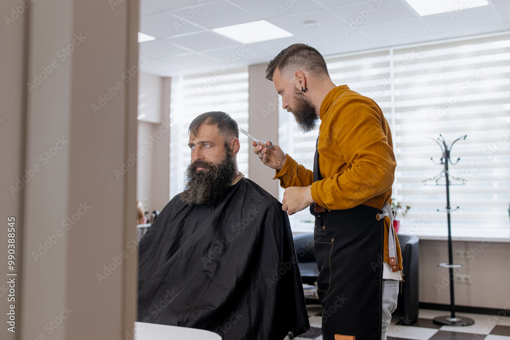 Stylish handsome barber in orange clothes and apron doing haircut to man and trimming beard in barber shop Stylish handsome barber in orange clothes and apron doing haircut to man and trimming beard in barber shop