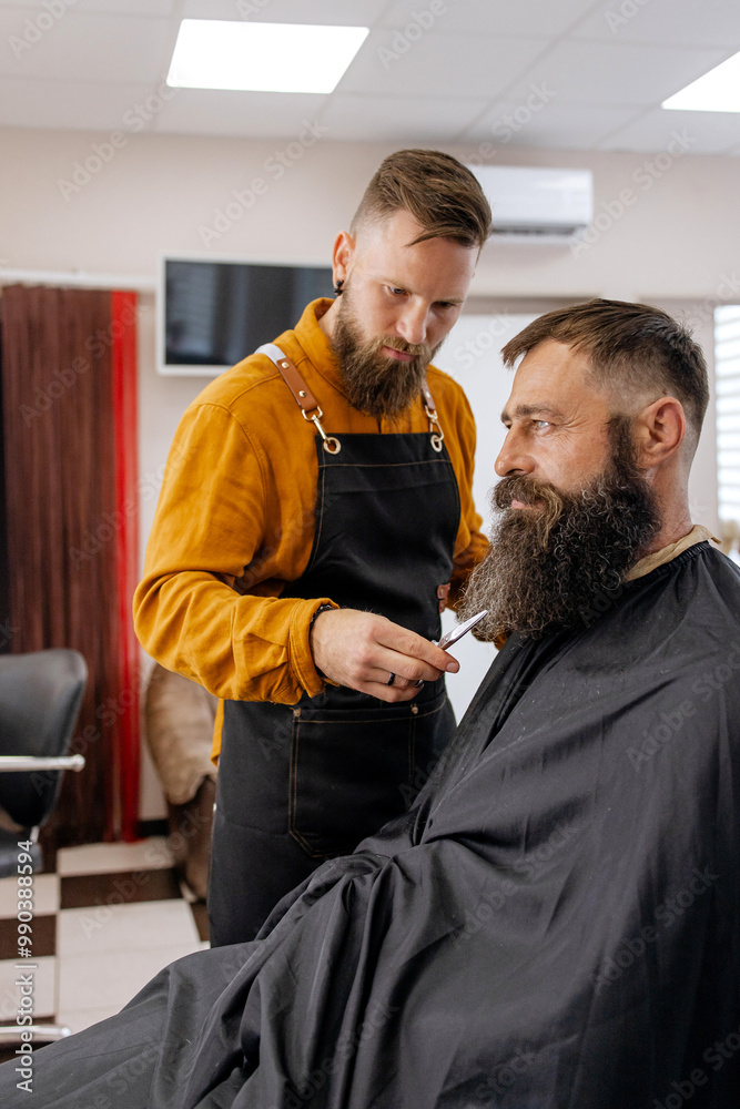 Stylish handsome barber in orange clothes and apron doing haircut to man and trimming beard in barber shop Stylish handsome barber in orange clothes and apron doing haircut to man and trimming beard in barber shop