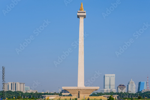 The image shows Monas (National Monument) in Jakarta, Indonesia, against a clear blue sky. The tall, white obelisk is topped with a golden flame, symbolizing the nation's independence and spirit.