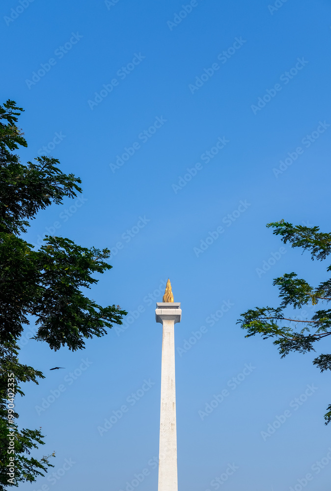 Monas (National Monument) in Jakarta, Indonesia, against a clear blue sky with trees and birds. The tall, white obelisk is topped with a golden flame, symbolizing the nation's independence and spirit.