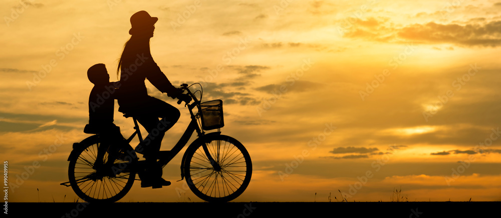 Obraz premium Silhouette of a mother and child riding a bicycle at sunset. Outdoor leisure activity with a beautiful sky background. Happy Family bonding and healthy lifestyle concept-banner image.
