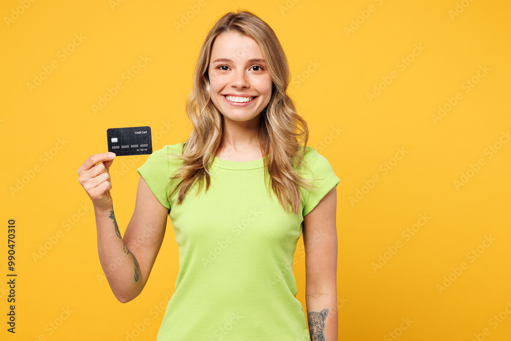 Young smiling cheerful happy blonde woman she wear green t-shirt casual clothes hold in hand mock up of credit bank card isolated on plain yellow orange background studio portrait. Lifestyle concept.
