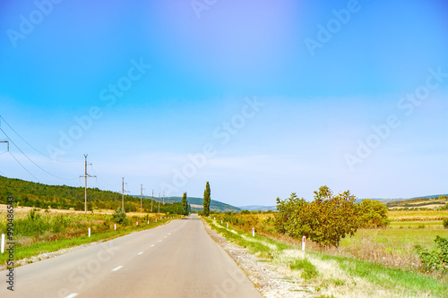 A long, straight rural road with power lines running alongside it, surrounded by fields and trees, under a clear blue sky.