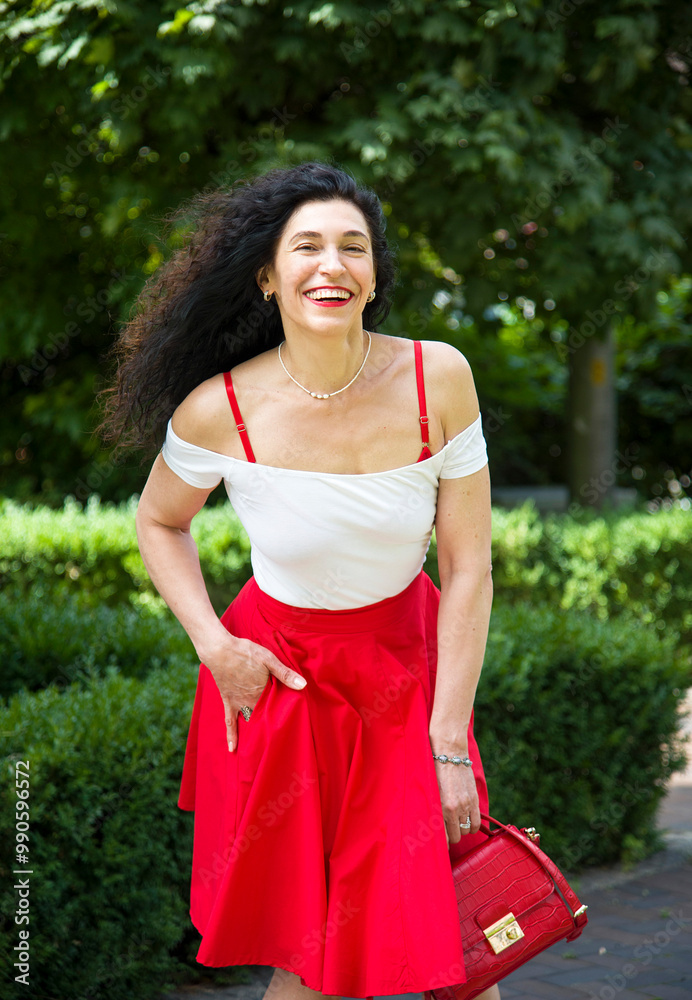 smiling brunette woman with curly hair and red lips in red skirt posing with red bag in park on sunny summer day smiling brunette woman with curly hair and red lips in red skirt posing with red bag in park on sunny summer day