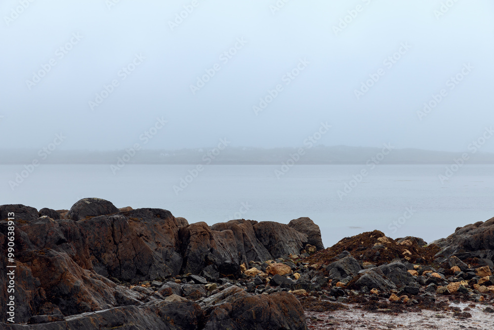 A misty coastal scene at Coral Strand Beach in Connemara, Ireland, with dark, rugged rocks and calm waters fading into the fog, creating a peaceful and secluded atmosphere under an overcast sky