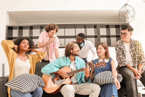 A young, handsome man is happily playing acoustic guitar on a sofa in a domestic house, surrounded by multiracial friends who are singing a song together and sharing a warm moment of togetherness