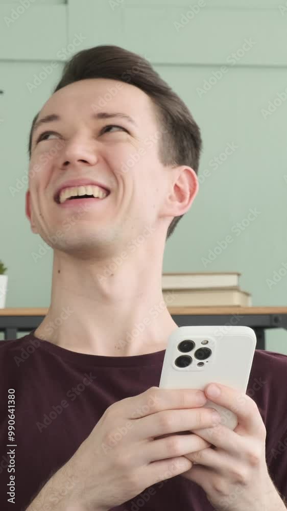 Joyful young adult Caucasian man messaging online using mobile phone and laughing, spending leisure time on sofa at home. Vertical video. Joyful young adult Caucasian man messaging online using mobile phone and laughing, spending leisure time on sofa at home. Vertical video.