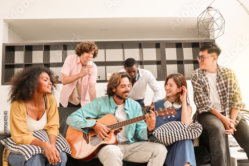 A young, handsome man is happily playing acoustic guitar on a sofa in a domestic house, surrounded by multiracial friends who are singing a song together and sharing a warm moment of togetherness