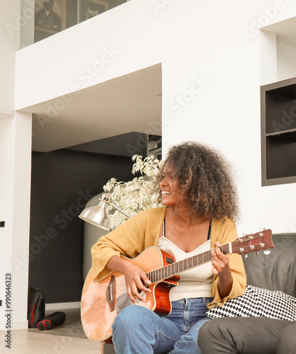 A happy, talented Black mixed-race woman is playing acoustic guitar and singing a song on a sofa in a domestic house. A beautiful female artist is practicing her singing skills before a performance.