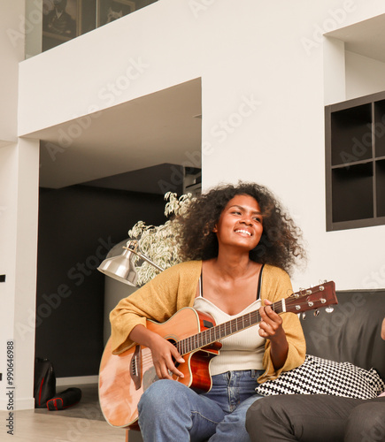A happy, talented Black mixed-race woman is playing acoustic guitar and singing a song on a sofa in a domestic house. A beautiful female artist is practicing her singing skills before a performance.