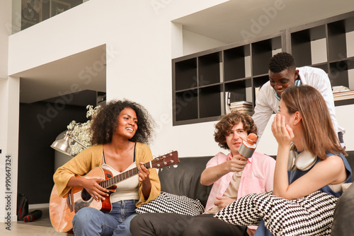 A young Black woman is happily playing acoustic guitar on a sofa in a domestic house, surrounded by multiracial friends who are singing a song together and sharing a warm moment of togetherness.