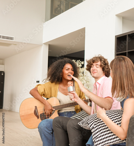 Happy mixed-race teenagers playing acoustic guitar on a sofa in a domestic house. Three multiracial friends are singing a song together and sharing a warm moment of togetherness.