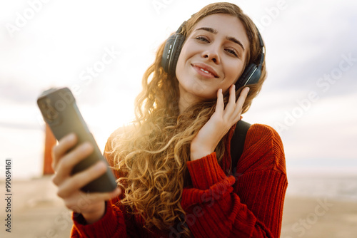 Young woman enjoying music by the waterfront while wearing headphones during a cloudy day by a lighthouse. Travel, tourism, technology, blogging concept. photo