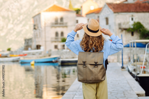 A young woman traveler enjoys the serene waterfront view in a coastal village during sunset time. Back view. Europe travel. Lifestyle, vacation, tourism, nature, active life.