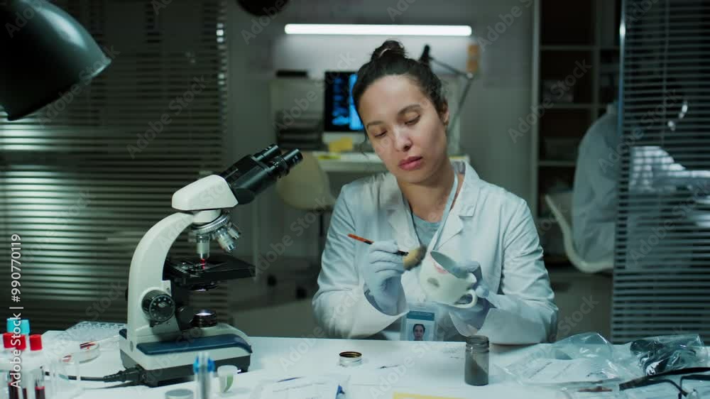 Female forensic scientist in gloves and white coat dusting mug for fingerprints with powder and brush while working in crime lab Female forensic scientist in gloves and white coat dusting mug for fingerprints with powder and brush while working in crime lab