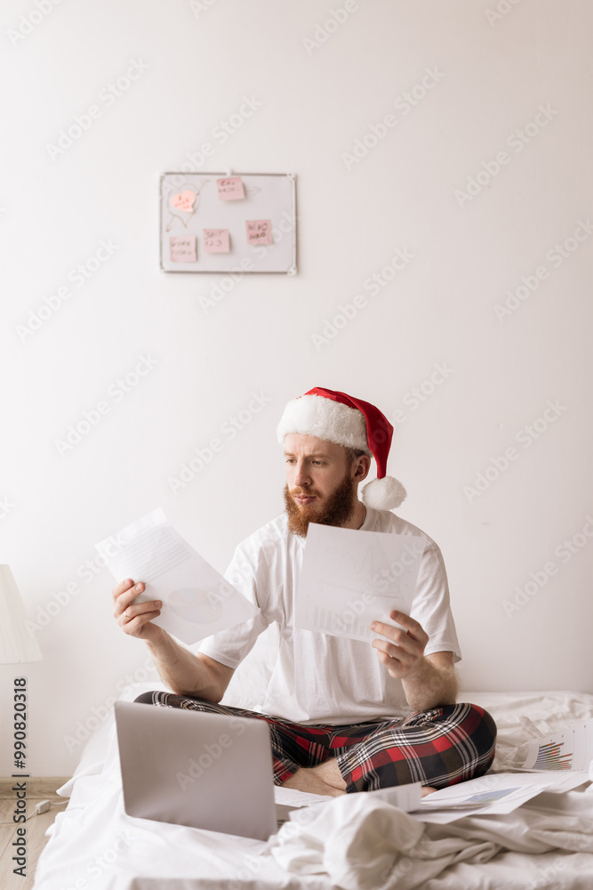 Concerned young man sitting in bed spending time in bedroom at home in Christmas morning. Remote work concept. Guy in Santa hat working with laptop and papers documents Concerned young man sitting in bed spending time in bedroom at home in Christmas morning. Remote work concept. Guy in Santa hat working with laptop and papers documents