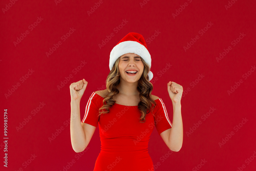 Shocked young brunette Santa woman in dress Christmas hat doing winner gesture clenching fists isolated on red colour background, studio portrait. Happy New Year celebration merry holiday concept Shocked young brunette Santa woman in dress Christmas hat doing winner gesture clenching fists isolated on red colour background, studio portrait. Happy New Year celebration merry holiday concept
