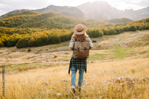 Back view.Travel woman explores a scenic mountain landscape during autumn in an open field with rolling hills and trees in the distance. Enjoying mountains view