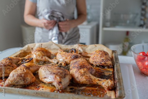 Woman with apron serving fresh and homemade cooked roast chicken legs on a baking tray in the kitchen