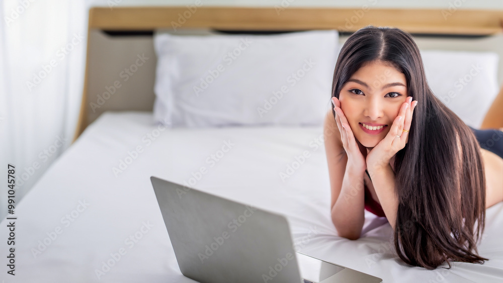 Fototapeta premium Pretty attractive sensuality young Asian woman wearing red bra and using laptop, laying on the white bed at bedroom in the morning