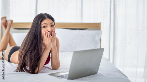 Pretty attractive sensuality young Asian woman wearing red bra and using laptop, laying on the white bed at bedroom in the morning