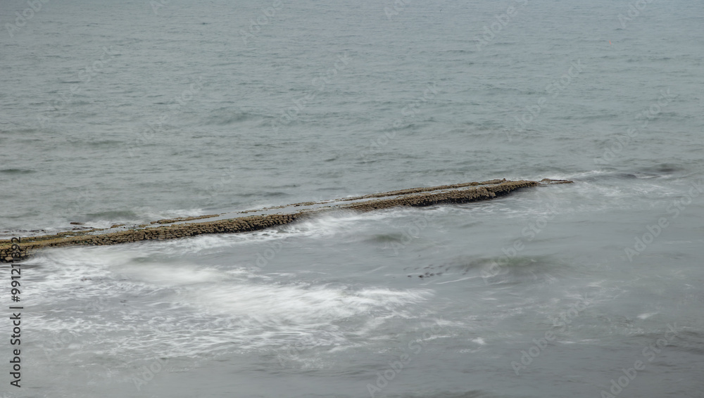 Fototapeta premium Long exposure of granite stone pier seawall and rocks along coast coastline in England, uk, Newcastle with blurred waves and grey water of the sea ocean