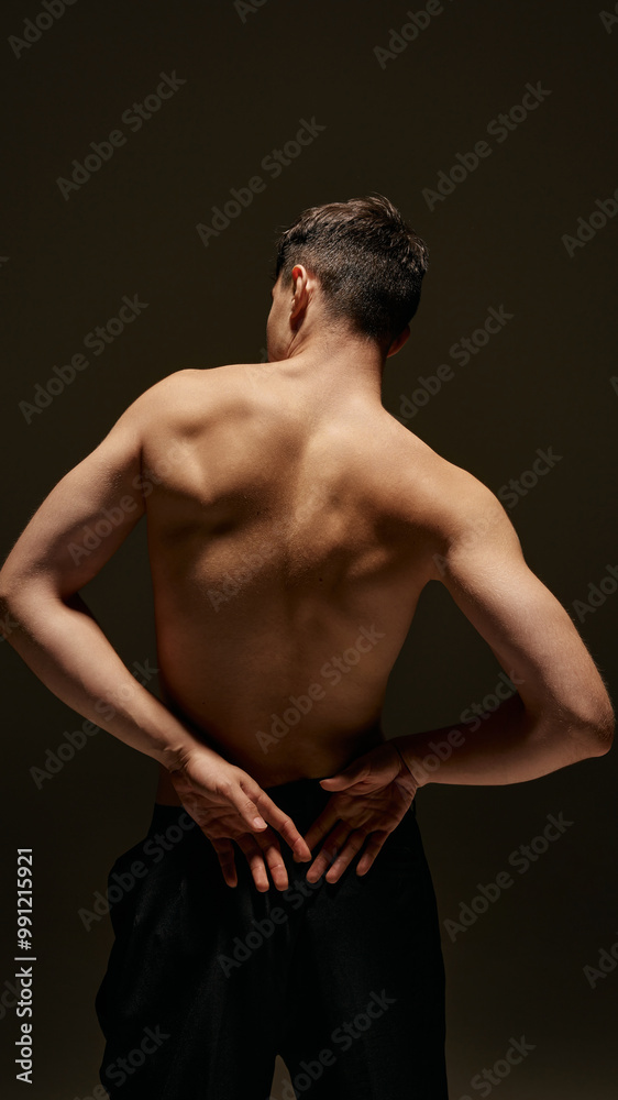 Fototapeta premium Rear view of young man posing shirtless in pants, showing his perfect, sportive body, relief strong back against dark studio background. Concept of male beauty, body and skin care, health, sport