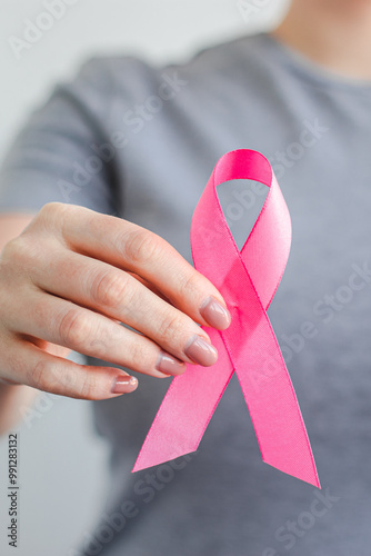 Woman in gray t-shirt holds pink ribbon in her hands. Breast Cancer Awareness Month. Supporting and promoting campaign against cancer. Health care concept. Vertical photo.