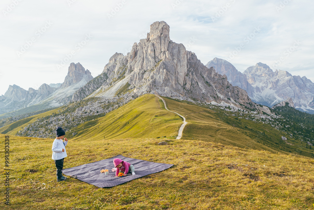 Naklejka premium Children Playing Outdoors on a Picnic Blanket in the Dolomites, Two young children playing on a picnic blanket in the Dolomites, surrounded by majestic mountain scenery and open nature.
