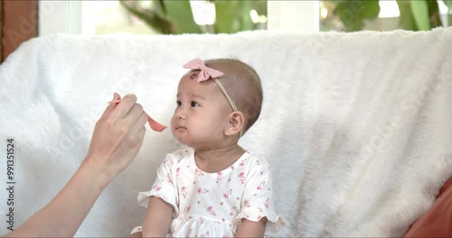 Cute and smiling Asian baby girl being fed with fruit puree. Candid shot – happy infant enjoys a meal. Child food, feeding equipment and utensils concept.