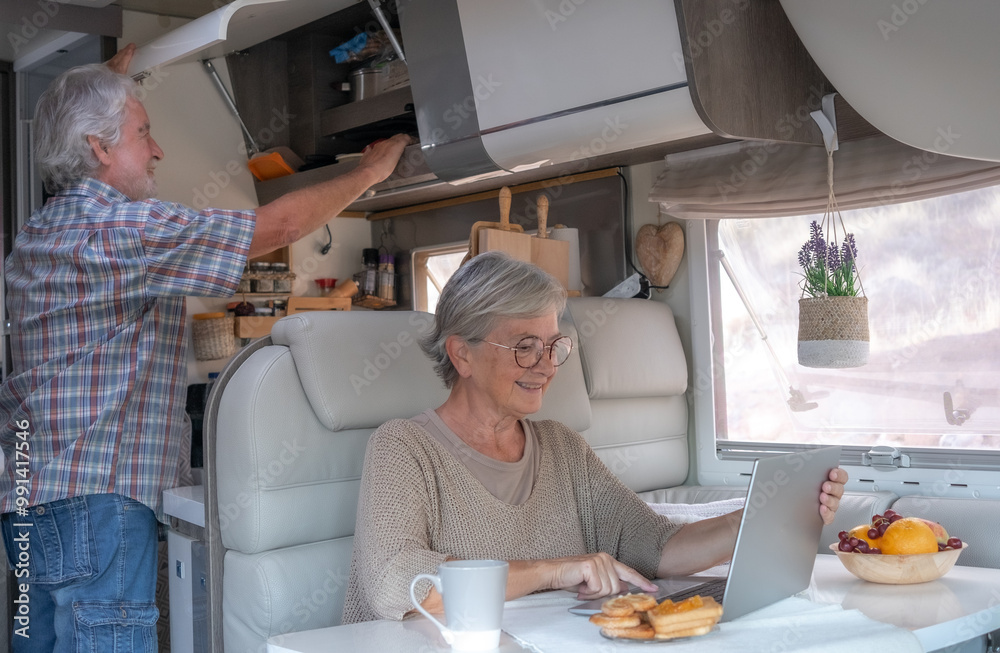 Relaxed senior couple in travel vacation leisure inside a camper van dinette enjoying breakfast together while wife uses laptop Relaxed senior couple in travel vacation leisure inside a camper van dinette enjoying breakfast together while wife uses laptop