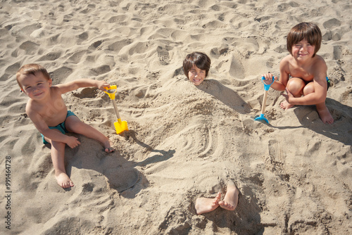 Fotografía Little brothers pose contentedly after burying their older brother in the sand on the beach with their shovels