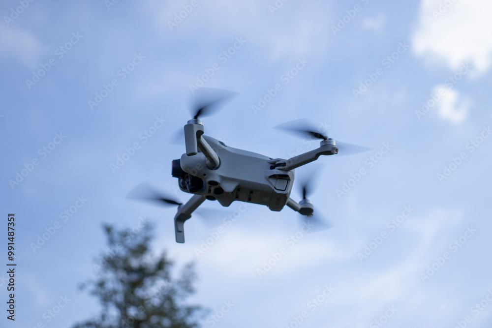 A flying drone captured mid-air against a clear blue sky with motion blur on the propellers. The background includes a blurred tree, suggesting the drone is being used outdoors. A flying drone captured mid-air against a clear blue sky with motion blur on the propellers. The background includes a blurred tree, suggesting the drone is being used outdoors.