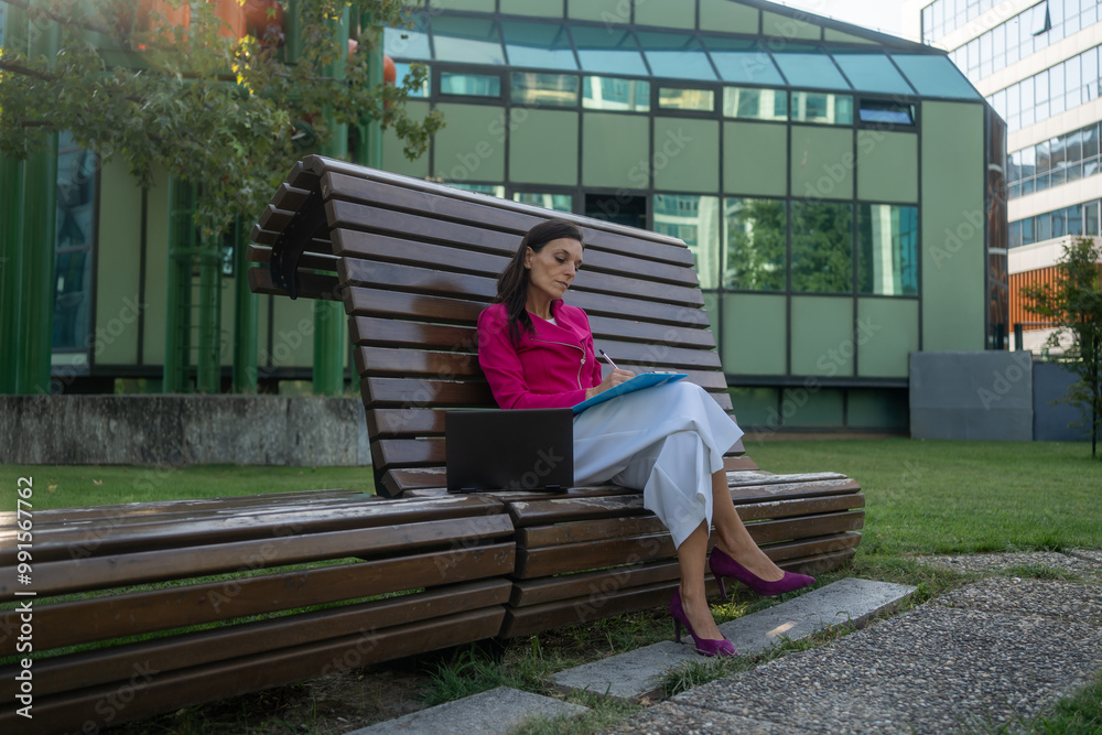 Businesswoman making notes looking at a laptop while sitting on bench in city park. Confident businesswoman in formal wear sitting in park in front of laptop and doing paperwork Businesswoman making notes looking at a laptop while sitting on bench in city park. Confident businesswoman in formal wear sitting in park in front of laptop and doing paperwork