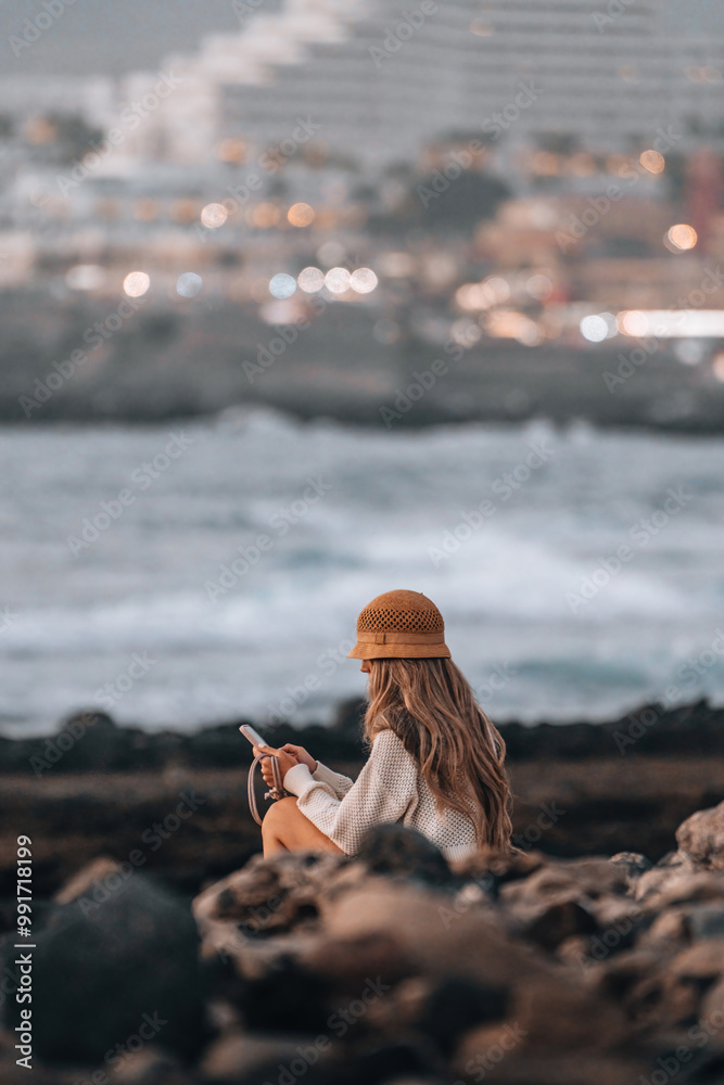 Obraz premium A woman is sitting on a rock by the ocean, looking at her phone