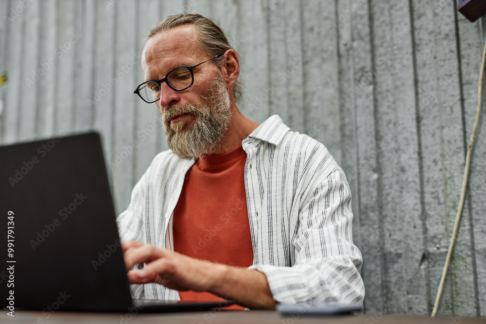 Minimal portrait of bearded adult man with suntan and glasses using laptop outdoors while sitting at table with metal wall in background copy space Minimal portrait of bearded adult man with suntan and glasses using laptop outdoors while sitting at table with metal wall in background copy space