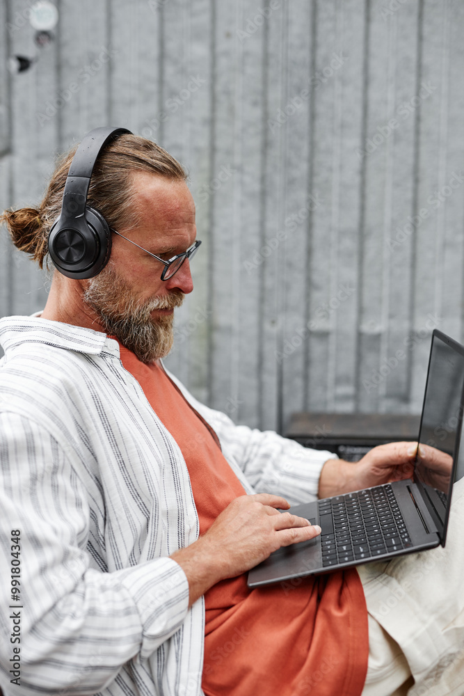 Vertical side view portrait of contemporary bearded man using laptop and wearing big headphones while working in outdoor setting with metal wall Vertical side view portrait of contemporary bearded man using laptop and wearing big headphones while working in outdoor setting with metal wall