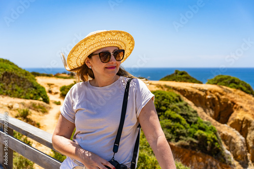 Summer vacation in Portugal. Portrait of woman in beige t-shirt, sunglasses and sun hat  standing on wooden pier on cliffs during sightseeing Algarve beaches on beautiful sunny day.