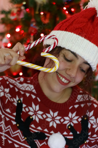 Smiling middle aged woman wearing a red and white Christmas-themed sweater with a reindeer design and a matching Santa hat. They are holding a candy cane near their mouth, which is pixelated for priva
