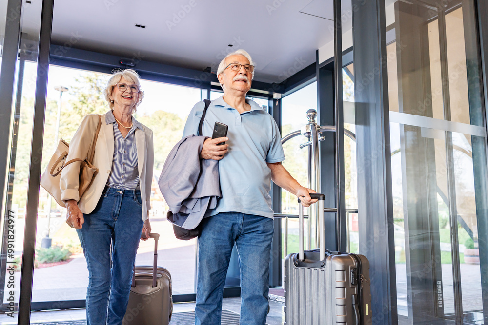 An elderly couple enters a hotel lobby, pulling their suitcases. They are dressed casually, smiling, and ready for travel, suggesting they are on a vacation or trip. An elderly couple enters a hotel lobby, pulling their suitcases. They are dressed casually, smiling, and ready for travel, suggesting they are on a vacation or trip.