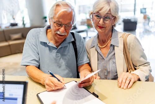 A smiling elderly couple stands in front of a hotel desk, filling out a check-in form or signing a bill, indicating they are either checking in or checking out after a wonderful stay.