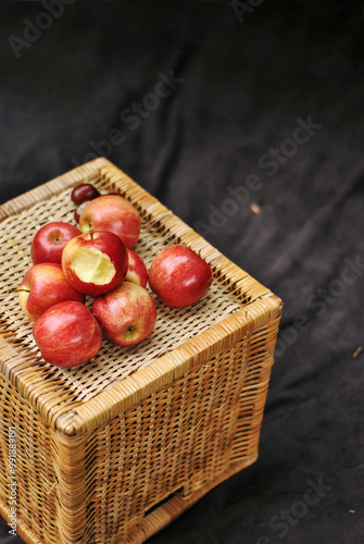 A bunch of red apples on the top of a wicker basket on the brown blanket. One of the apples is bitten off.