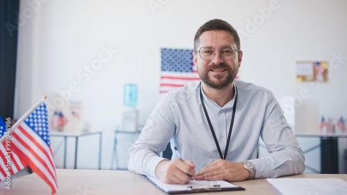 Camera moving closer to Caucasian male with beard and glasses sitting at desk. Finishing filling information on piece of paper. Using pen to write information. American flags in background.