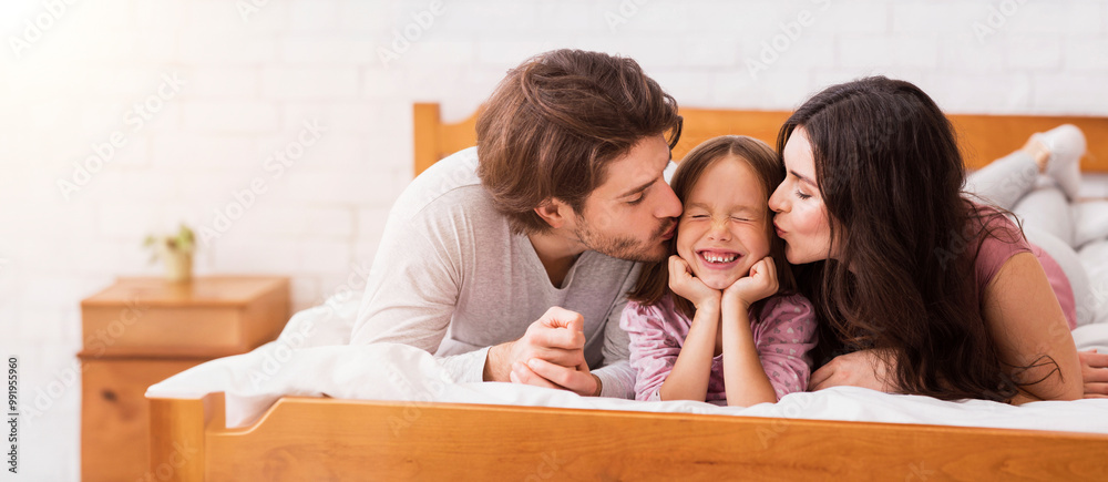 Parents Love. Portrait Of Loving Mother And Father Giving A Kiss To Their Little Daughter From Both Sides While Lying Together In Bed Parents Love. Portrait Of Loving Mother And Father Giving A Kiss To Their Little Daughter From Both Sides While Lying Together In Bed