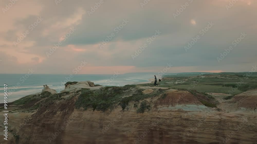 Female surfer and her dog running on the hills by the sea at beautiful pink sunset. Woman with surfboard running after the sunset surf session on the coastline. Female surfer and her dog running on the hills by the sea at beautiful pink sunset. Woman with surfboard running after the sunset surf session on the coastline.
