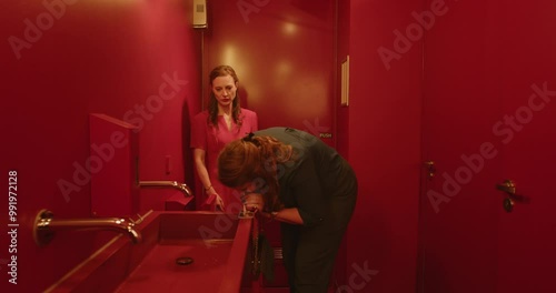 Two woman doing drugs in a public bathroom. One woman instructs the other in how to snort cocaine from the mirror