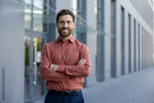 Confident male professional stands with arms crossed outside modern urban building, expressing confidence and professionalism. Businessman wearing casual attire represents successful entrepreneur