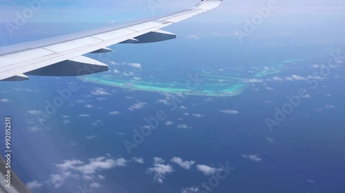 The view from the plane window of the ocean and the atoll. The silver wing of an airplane. There are clouds in places. Turquoise water and snow-white beaches of islands. The Indian Ocean and Maldives